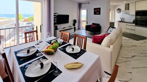 Uma sala de jantar com mesa posta para dois, sofá, televisão e cozinha ao fundo.