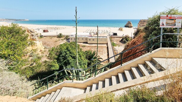 Vista da praia com uma escadaria descendo até a areia, um aviso e um penhasco rochoso.