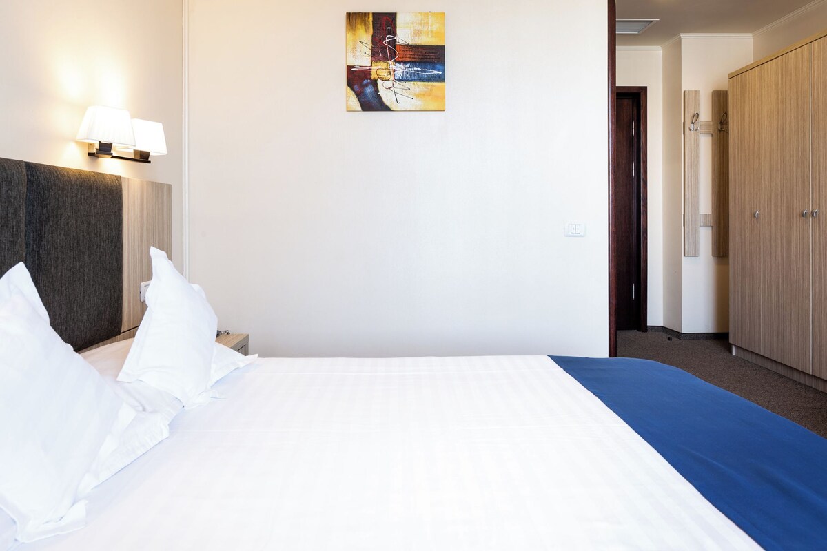 A hotel room with a bed, a nightstand, a lamp, a painting, and a wardrobe.