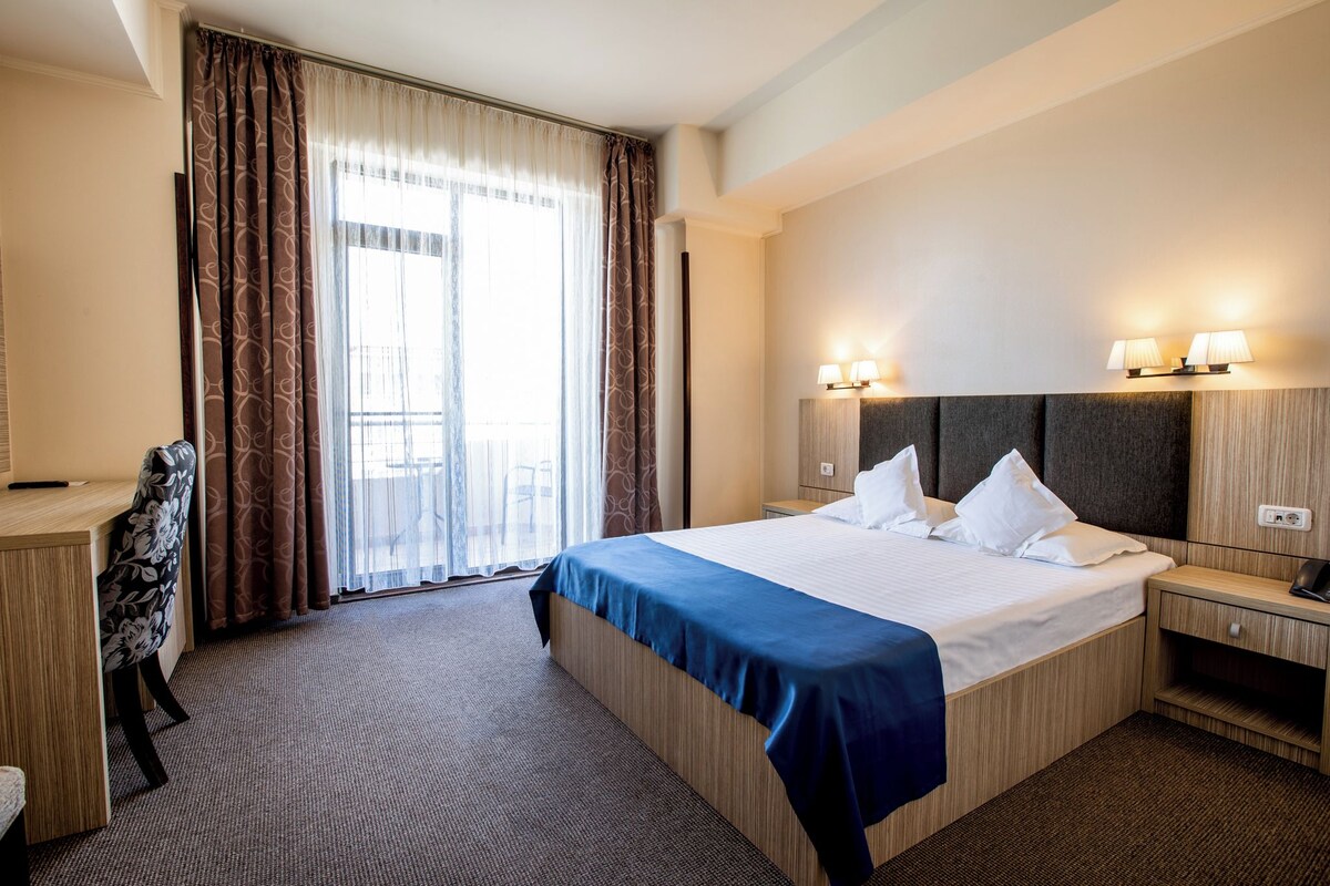 A hotel room with a large bed, a desk, a chair, and a window with curtains.