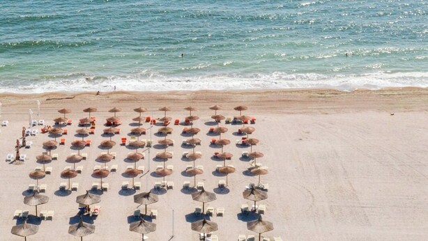 Uma praia com fileiras de espreguiçadeiras e guarda-sóis, areal e o oceano ao fundo.
