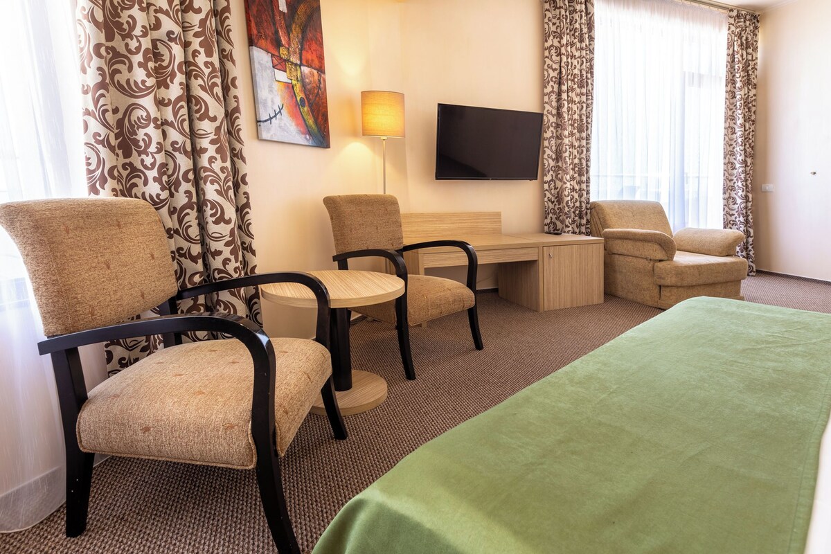 A hotel room with two chairs, a desk, a bed, a TV, and curtains.