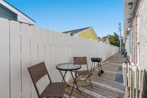 A deck with two wicker chairs and a glass table, a white picket fence, and a brick building with a yellow roof in the background.