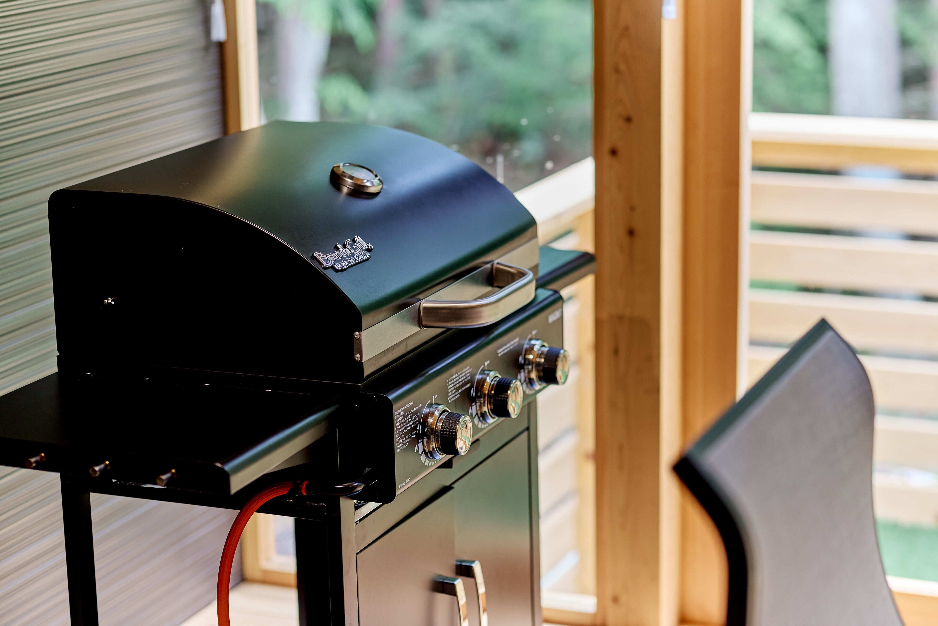 A black barbecue grill with a digital display and four control knobs, positioned in a wooden deck area with a view of greenery outside.