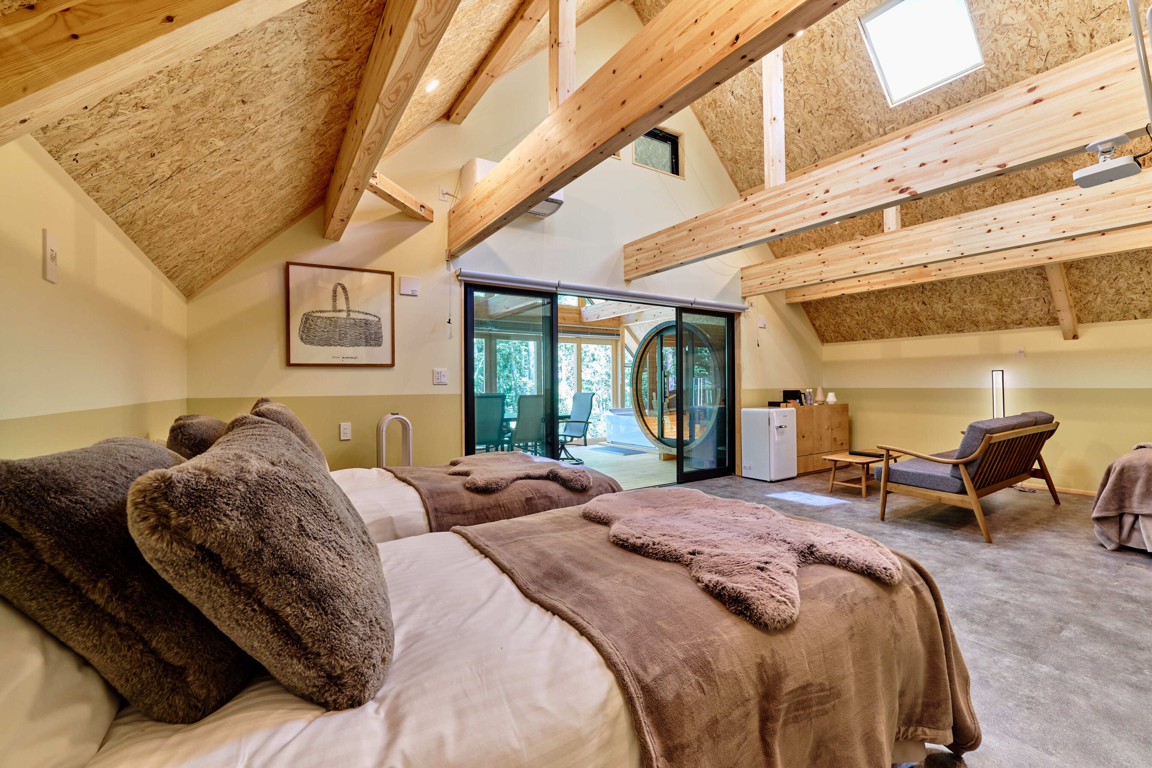 Ein gemütliches Zimmer mit Holzbalken, einem Bett mit brauner Bettwäsche, einem Sessel und Blick ins Freie.