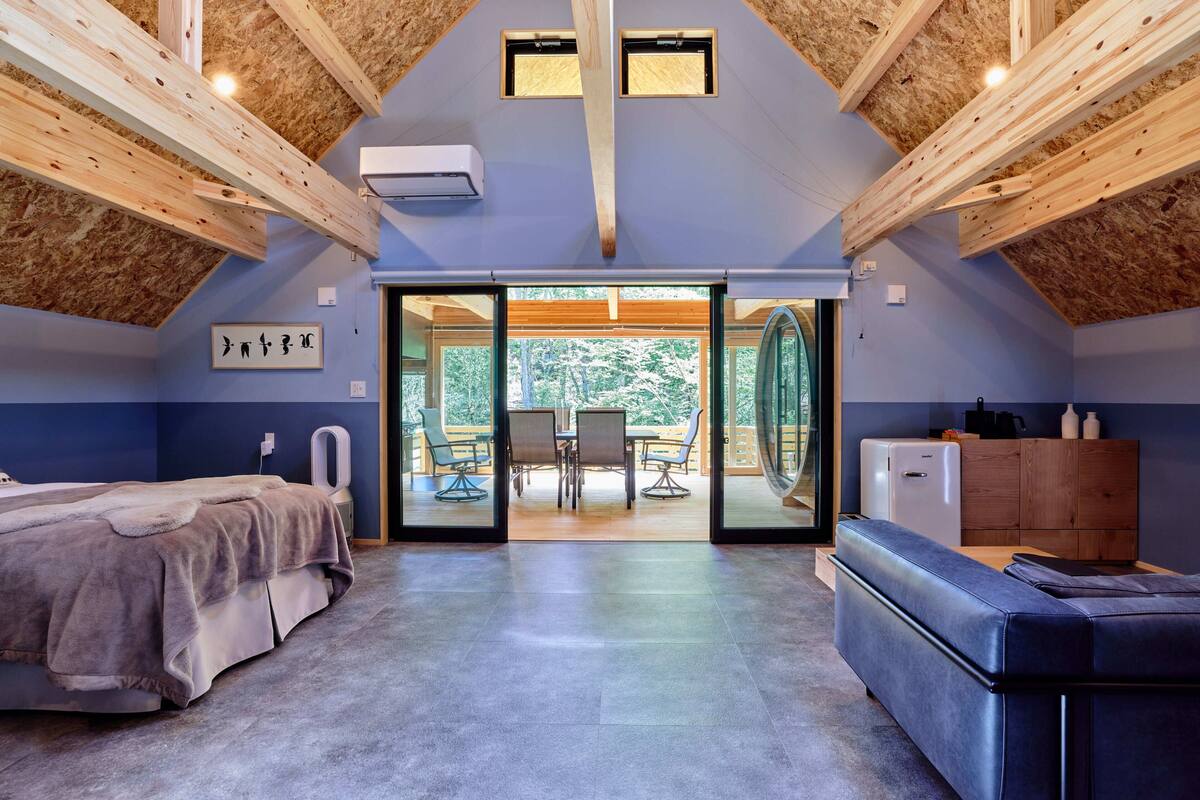 A modern bedroom with a bed, a sofa, a dining table with chairs, and a view of the outdoors.