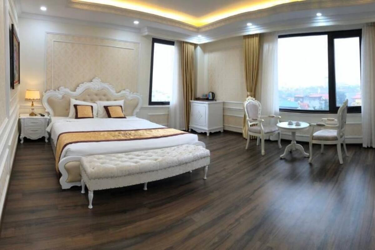 Ruime hotelkamer met een groot bed, een zithoek met een tafel en stoelen, en uitzicht op de stad door grote ramen.