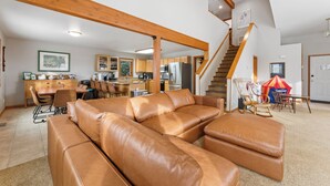 A spacious living area with a large leather sectional sofa, wooden beams on the ceiling, a kitchen in the background, and a staircase leading to another level.