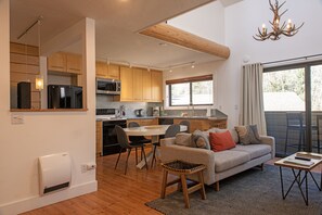 A modern kitchen with wooden cabinets, a dining area with a table and chairs, a living room with a sofa and a coffee table, and a large window with a view of the outdoors.