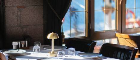 A dining area with a table set for two, a framed picture on the wall, and a view of the sea through large windows.