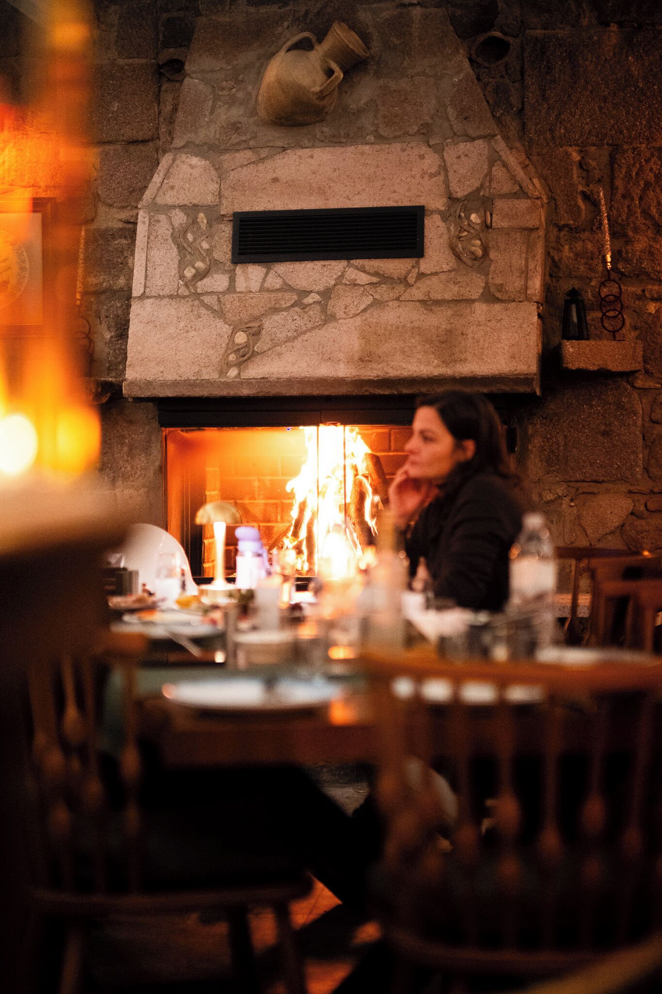 A stone fireplace with a vent above it, a table with various items, and a person sitting at the table.