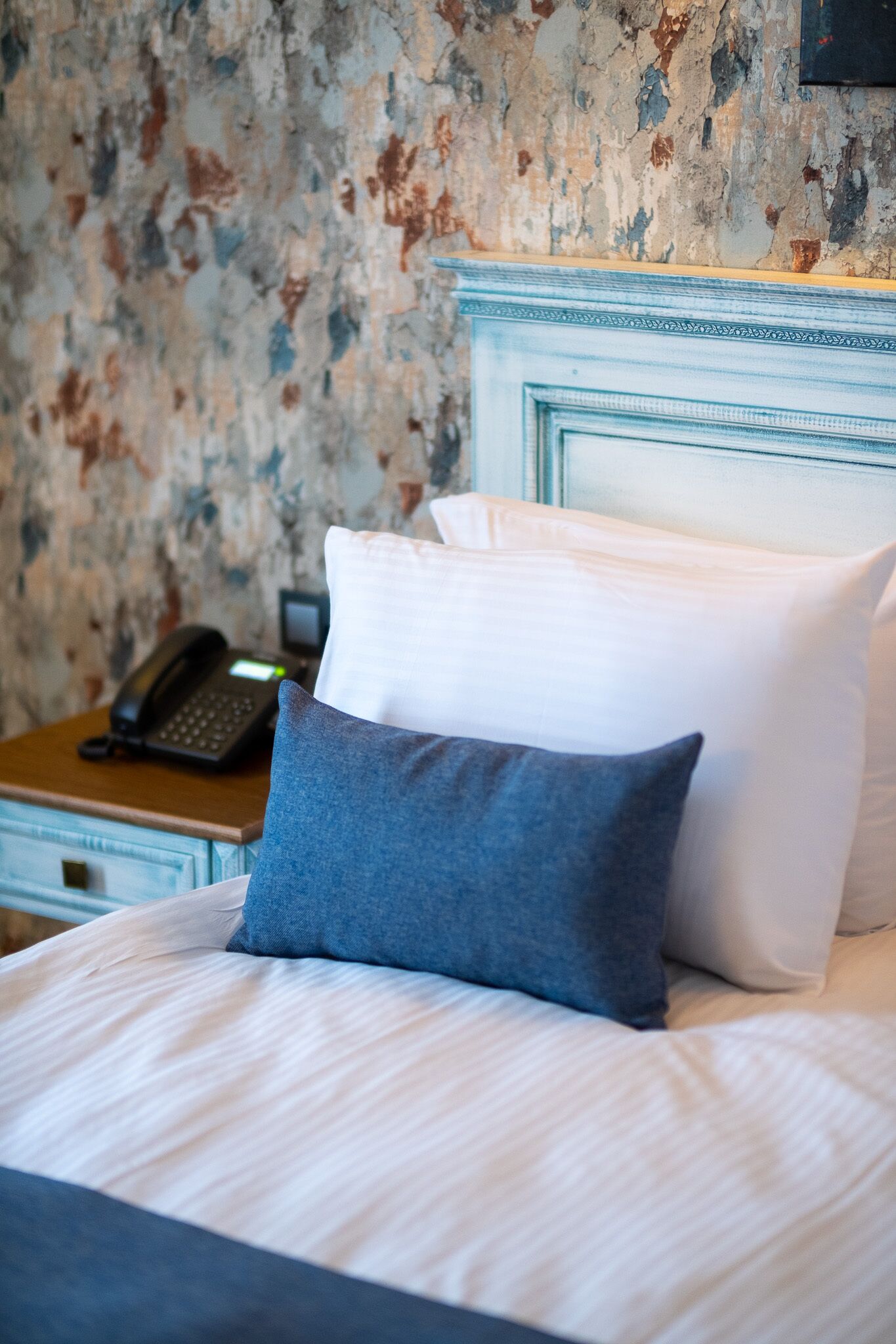 A neatly made bed with white and blue pillows, a wooden nightstand with a telephone, and a textured wall in the background.