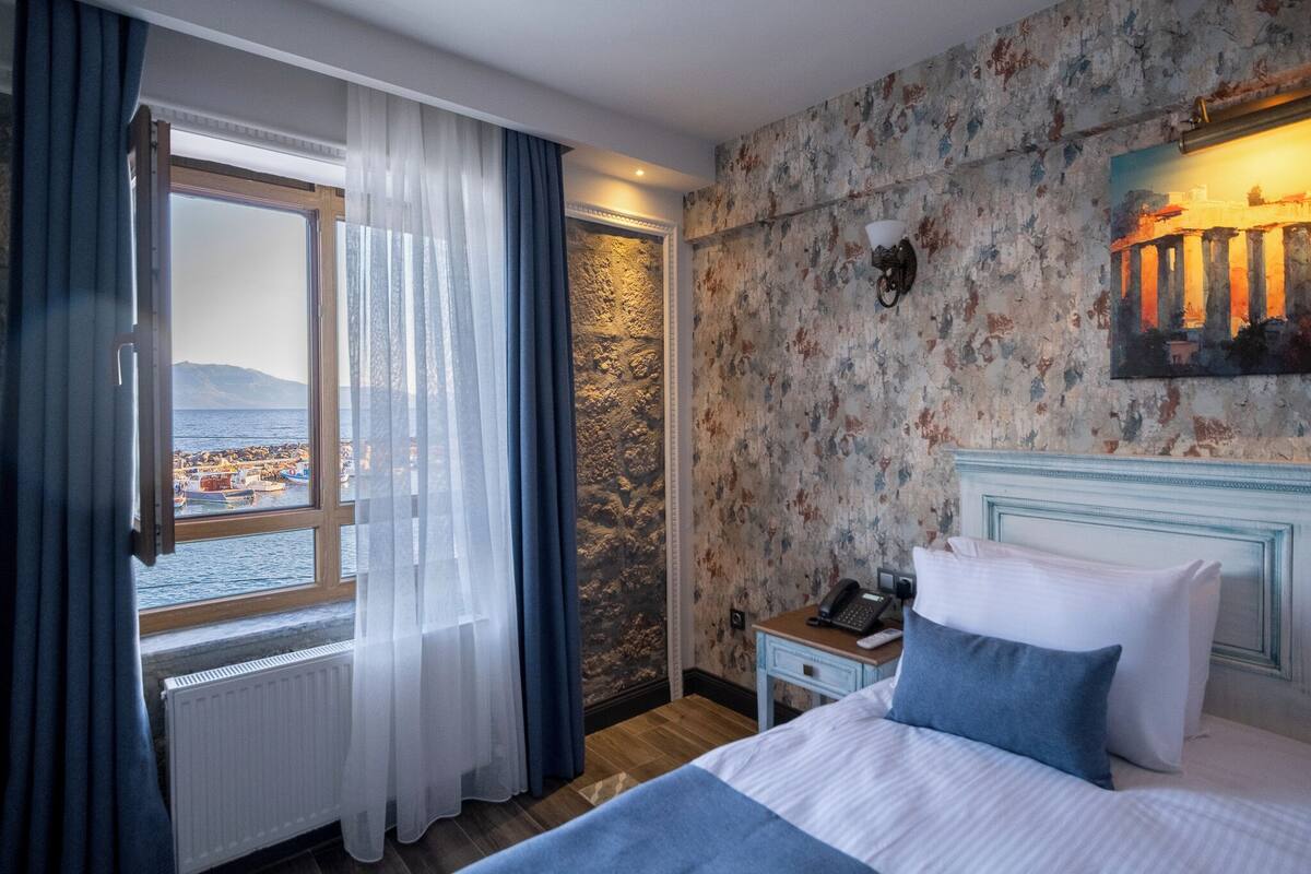 Une chambre d’hôtel avec un lit, une table de chevet, une lampe, une fenêtre avec des rideaux et une vue sur une ville côtière.