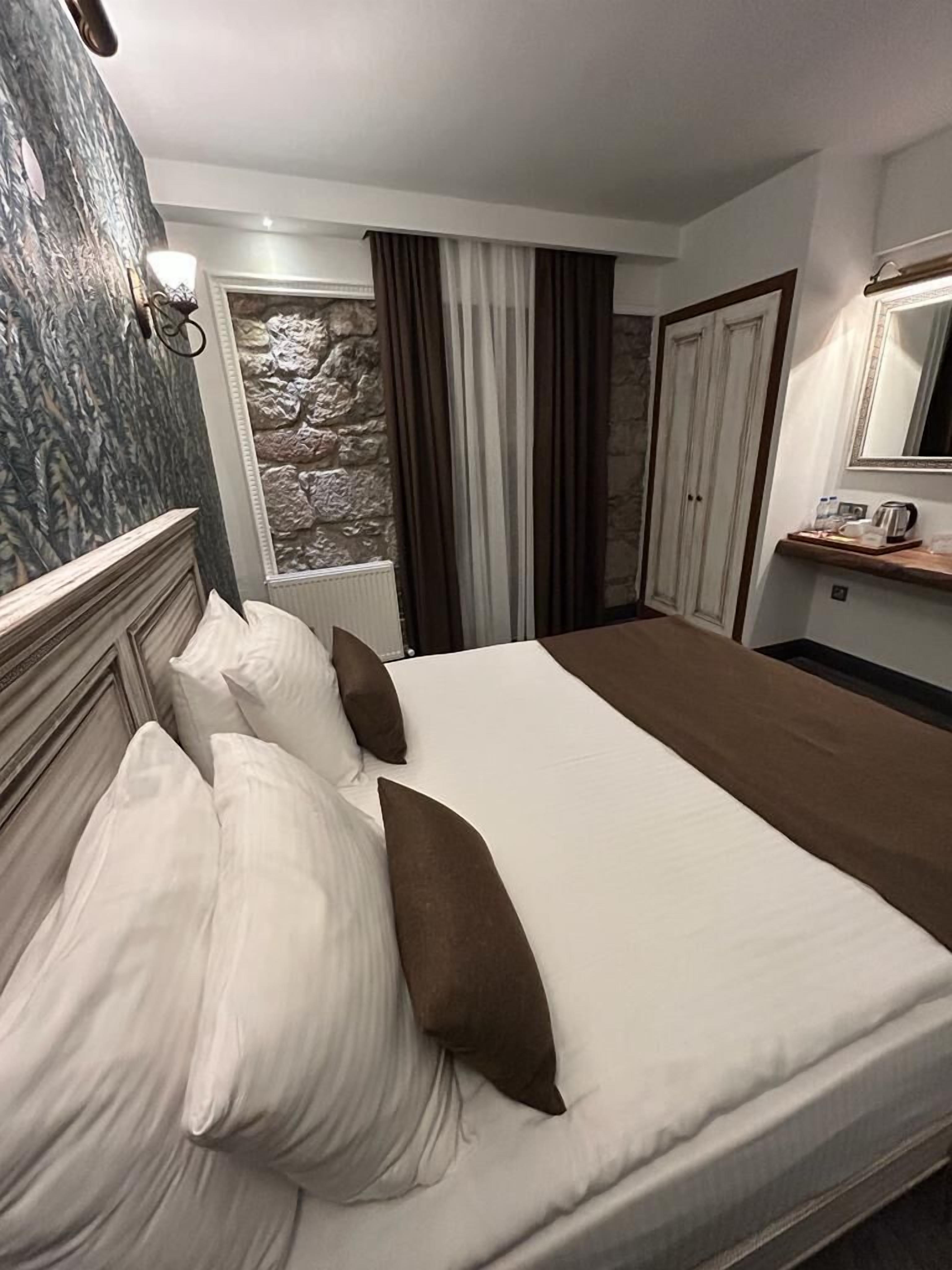 A hotel room with a large bed, a stone wall, a window with curtains, a wooden headboard, and a mirror above the bed.