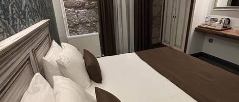 A hotel room with a large bed, a stone wall, a window with curtains, a wooden headboard, and a mirror above the bed.
