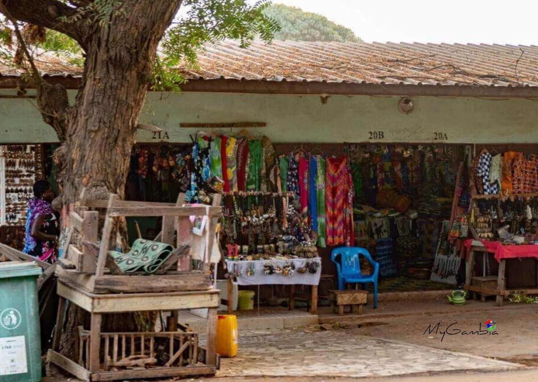 Un puesto de mercado con telas y joyas de colores, un árbol en primer plano y un edificio con indicaciones numeradas.