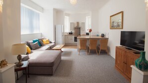 Ein modernes Wohnzimmer mit einer Couch, einem Hocker und einem Essbereich mit Tisch und Stühlen. Ein Fernseher steht auf einer Holzkommode, und im Hintergrund ist eine Küche zu sehen.