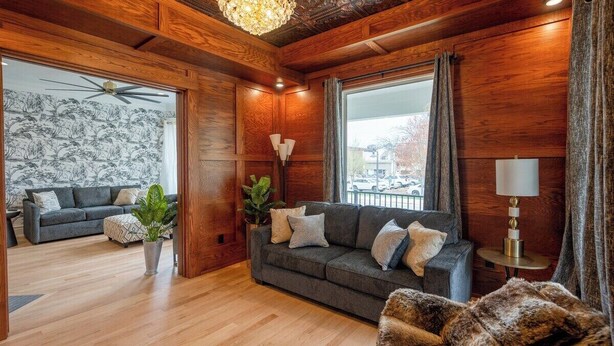 Ein gemütliches Wohnzimmer mit Holzwänden, einem grauen Sofa, einem Fellstuhl und einem großen Fenster mit Vorhängen.