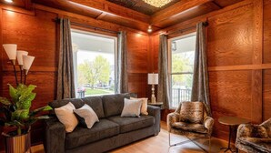 Ein gemütliches Wohnzimmer mit einem dunkelgrauen Sofa, zwei Sesseln und einem kleinen Tisch. Große Fenster mit Vorhängen lassen natürliches Licht und Ausblicke ins Freie herein.