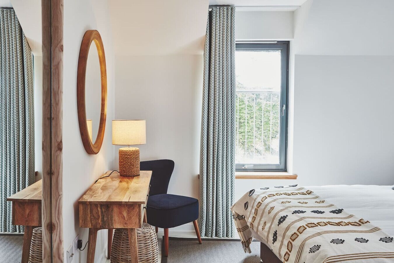 Ett hotellrum med ett skrivbord i trä, en stol, en säng med mönstrat sänglinne, en stor spegel och ett fönster med gardiner.