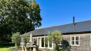 Ein Holzhaus mit Schornstein, eine Terrasse mit Korbmöbeln und ein großer Baum im Hintergrund.