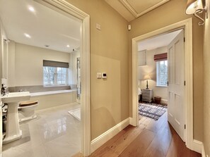 Une salle de bain avec un lavabo, des toilettes et une baignoire, un couloir avec un tapis, et une porte donnant sur une autre pièce.