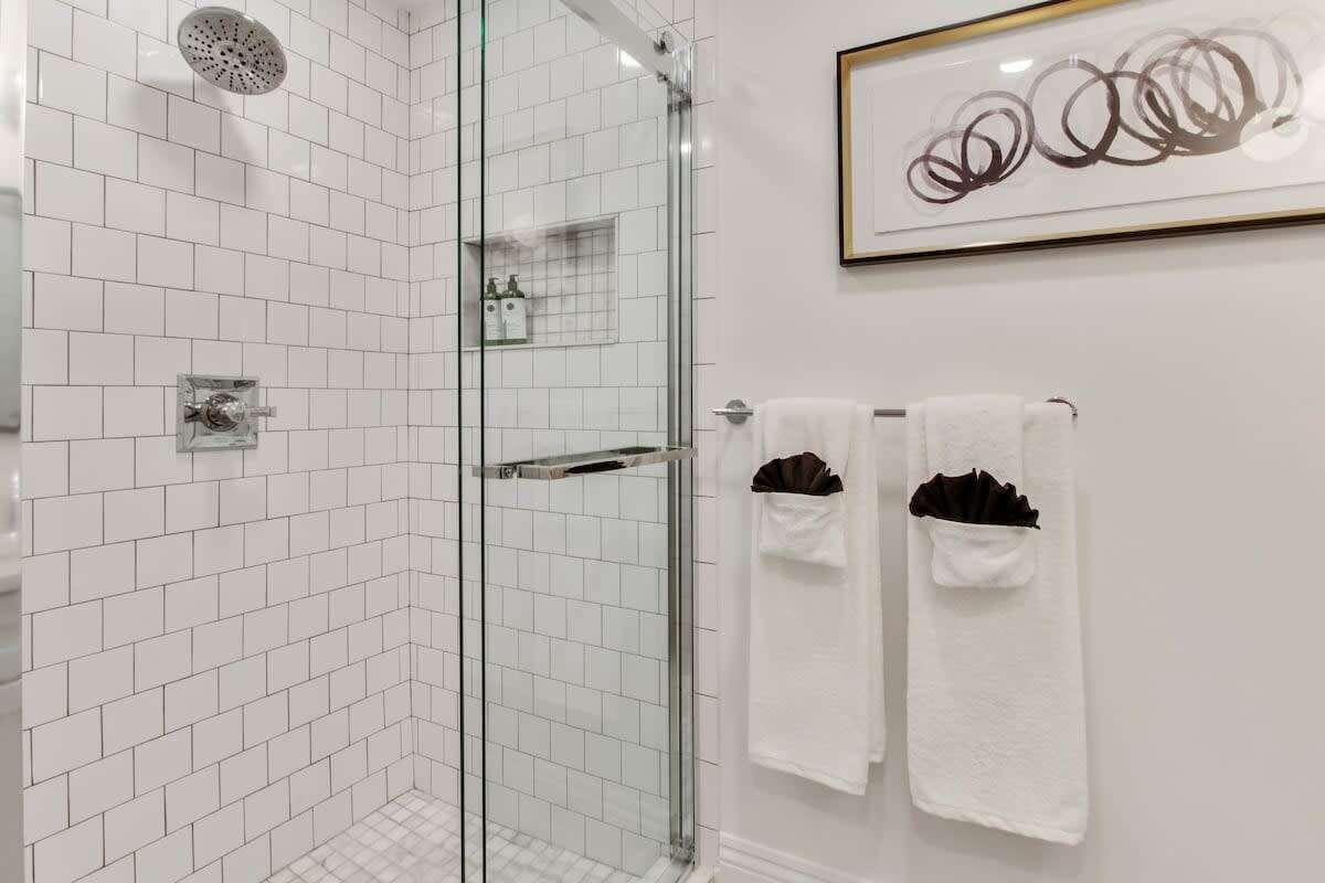 A modern bathroom with white tiled walls, a glass shower enclosure, and two white towels with brown accents.