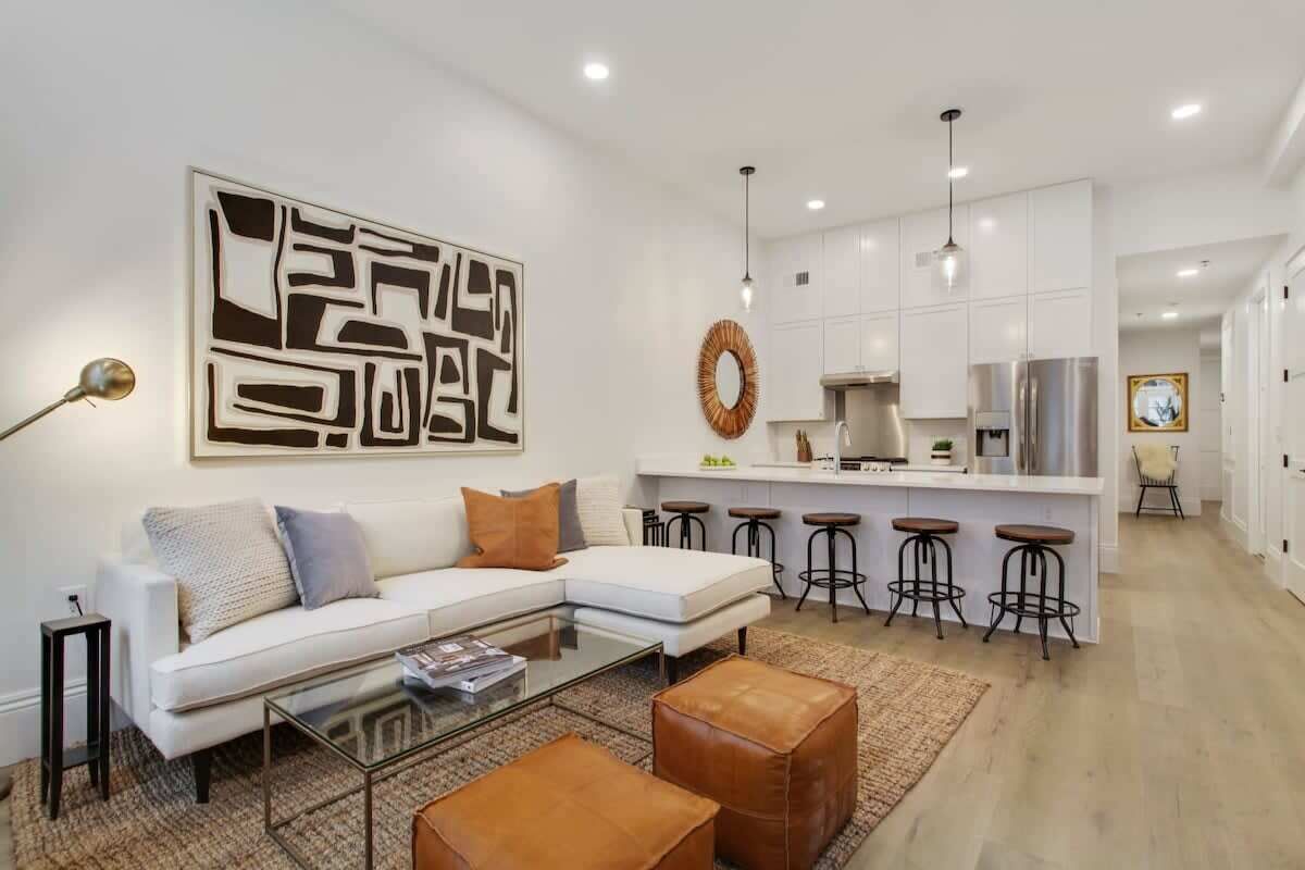 A modern living room with a white sofa, a glass coffee table, and a kitchen area with white cabinets and a stainless steel refrigerator.