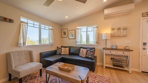 Un salon comprenant un canapé d’angle, un fauteuil, une table basse et un bureau avec une lampe. On y trouve également des fenêtres, un ventilateur de plafond et un appareil de climatisation.