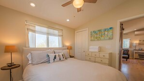 Une chambre avec un grand lit, des lampes de chevet, un ventilateur de plafond, une fenêtre avec des rideaux, une commode et un tableau au mur.