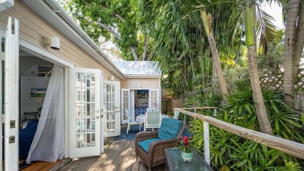 A wooden deck with wicker furniture and a small table, surrounded by lush greenery and palm trees.