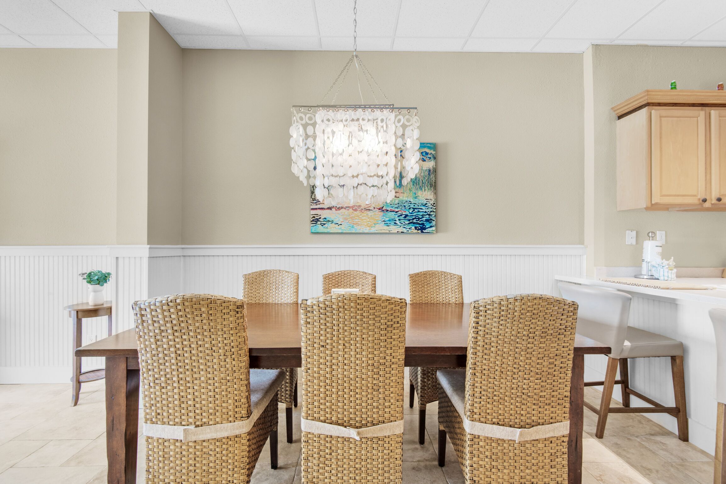 A dining area with a wooden table and wicker chairs, a hanging light fixture, and a painting on the wall.