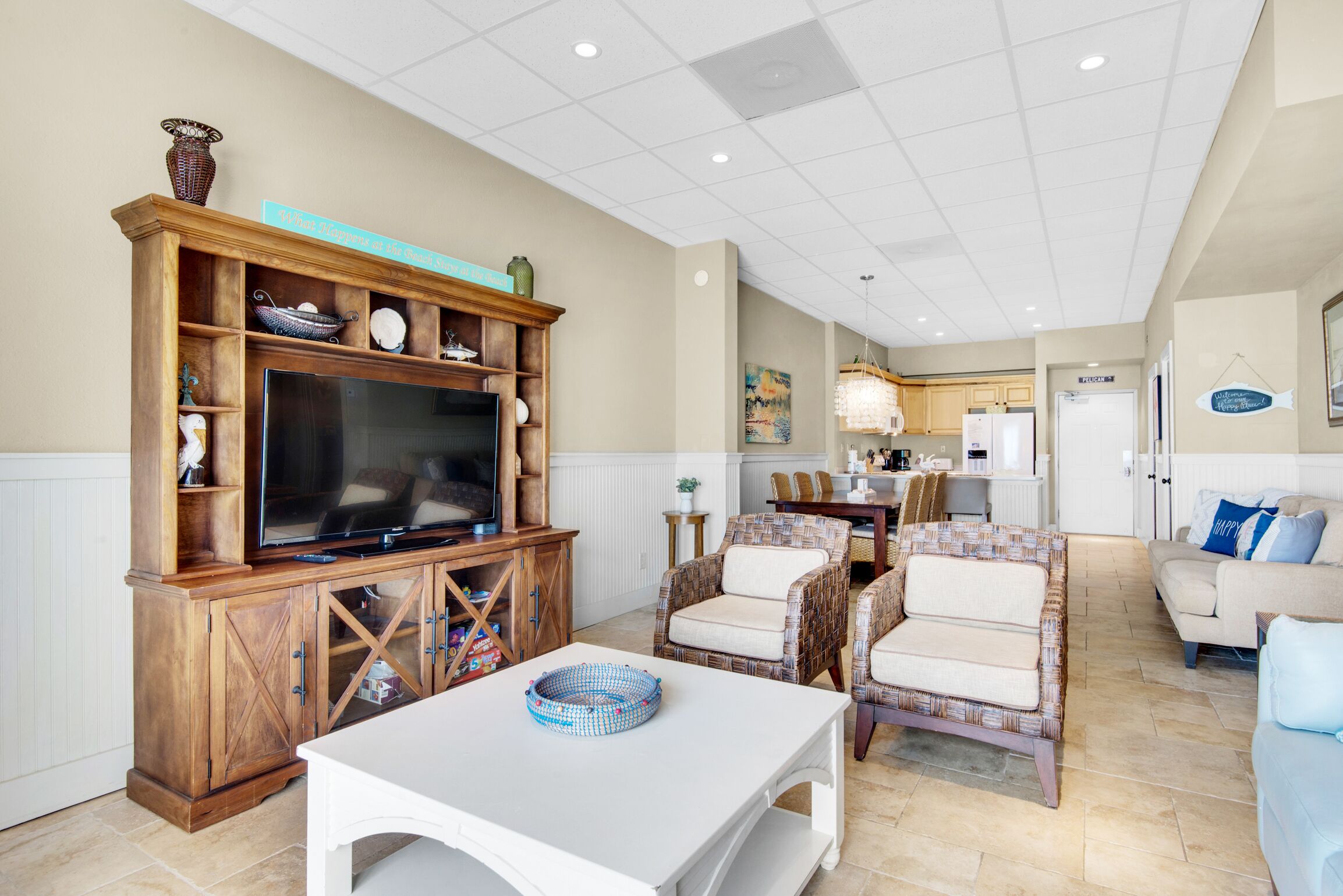 A living room with a television, wicker chairs, a white table, and a kitchen in the background.