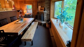 Een houten interieur met een lange eettafel, bankjes, een open haard en grote ramen.