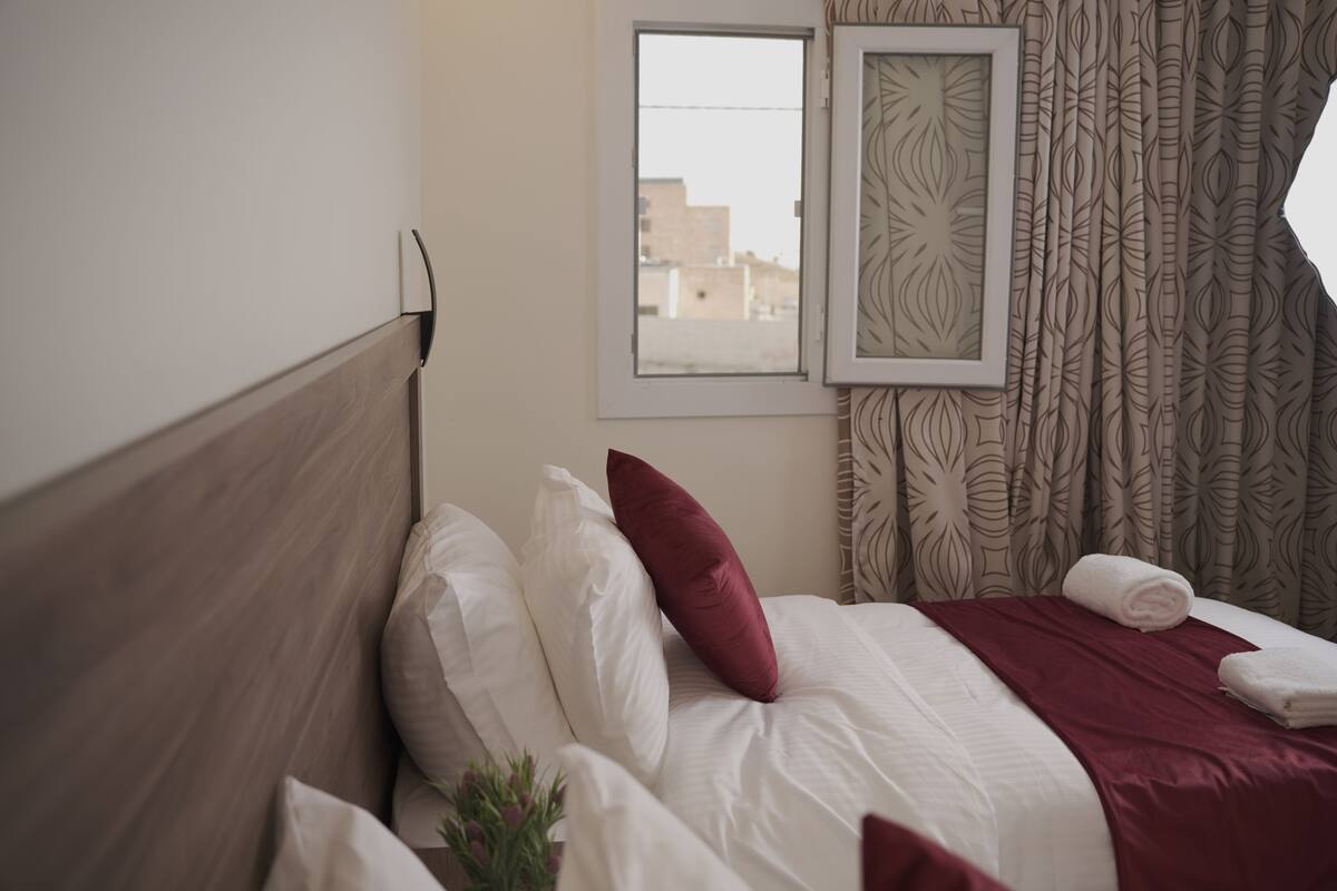 A neatly made bed with white and red pillows, a wooden headboard, and a window with patterned curtains.
