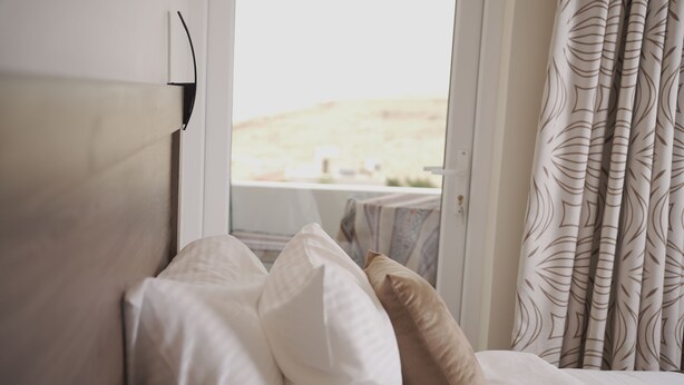 A bed with white and beige pillows, a window with a view of a landscape, and patterned curtains. - Petra Passion Inn (Wadi Musa)