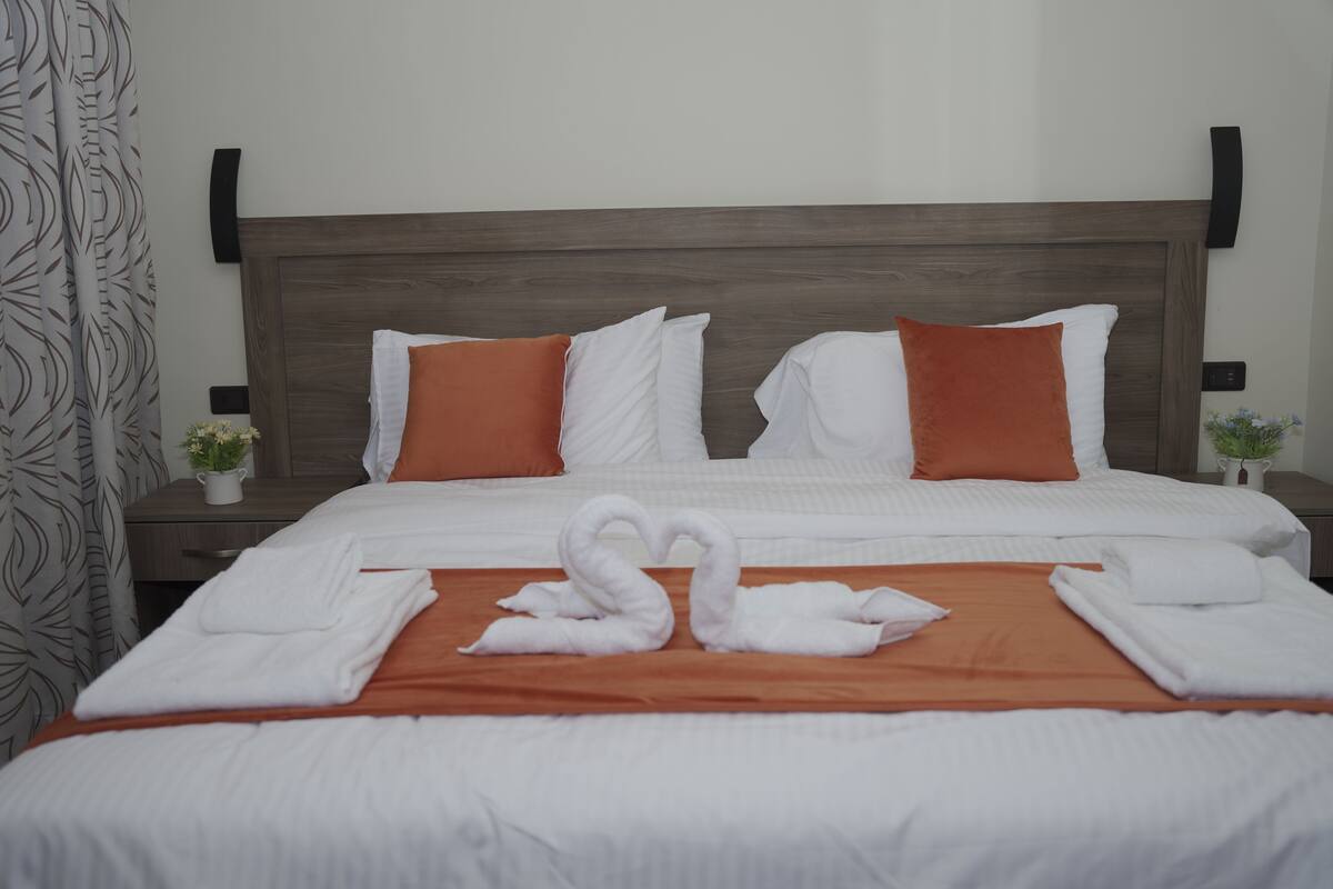 A neatly made bed with white and orange pillows, a brown headboard, and a swan-shaped towel arrangement.