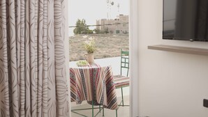 A hotel room with a bed, a TV mounted on the wall, a small table with a patterned tablecloth, a chair, and a view of the outside. - Petra Passion Inn (Wadi Musa)