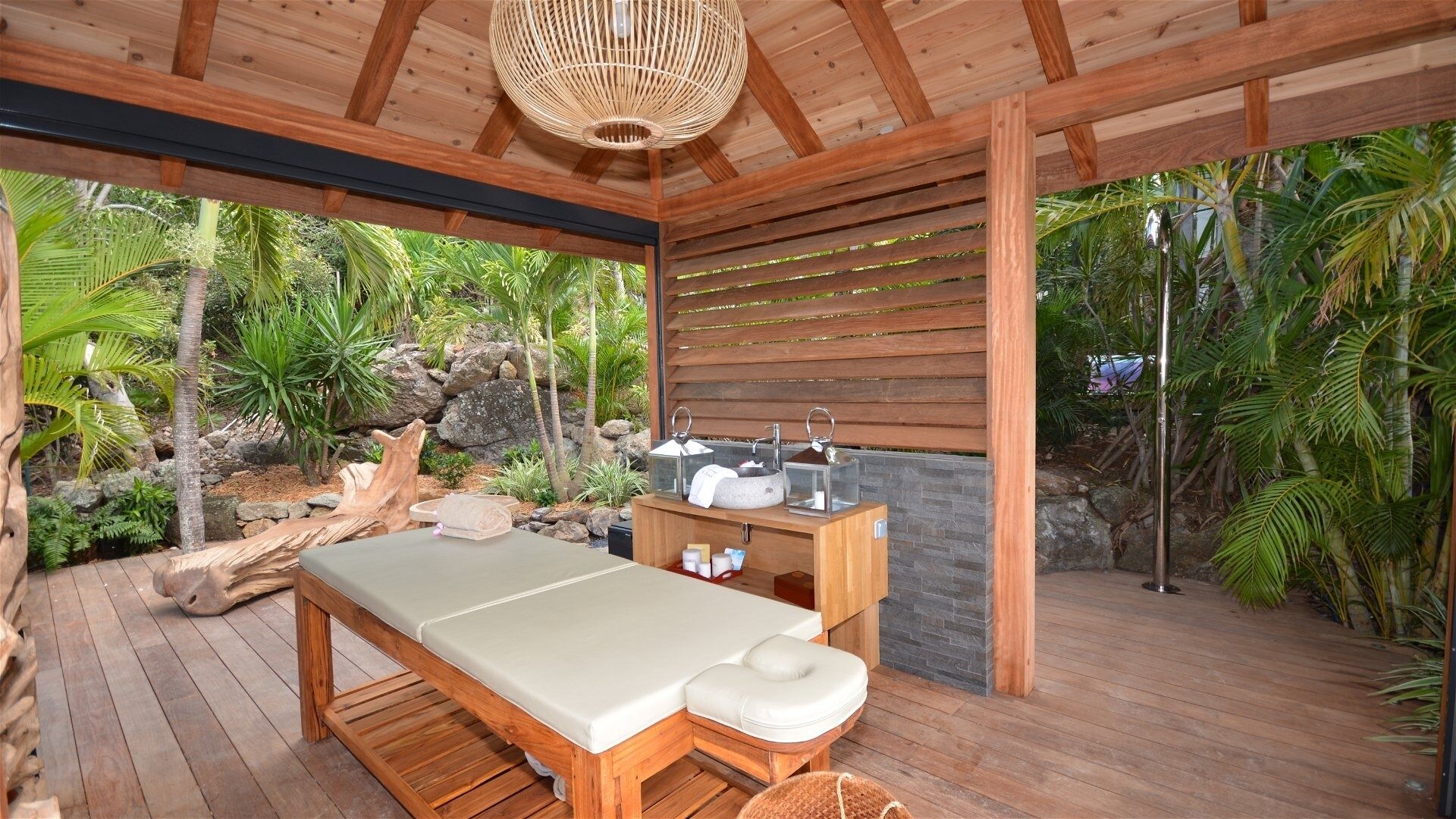 A wooden deck with a massage table, a stone wall, and a tropical garden view.