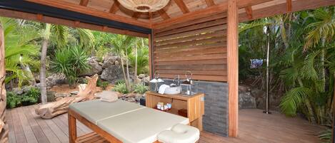 A wooden deck with a massage table, a stone wall, and a tropical garden view.