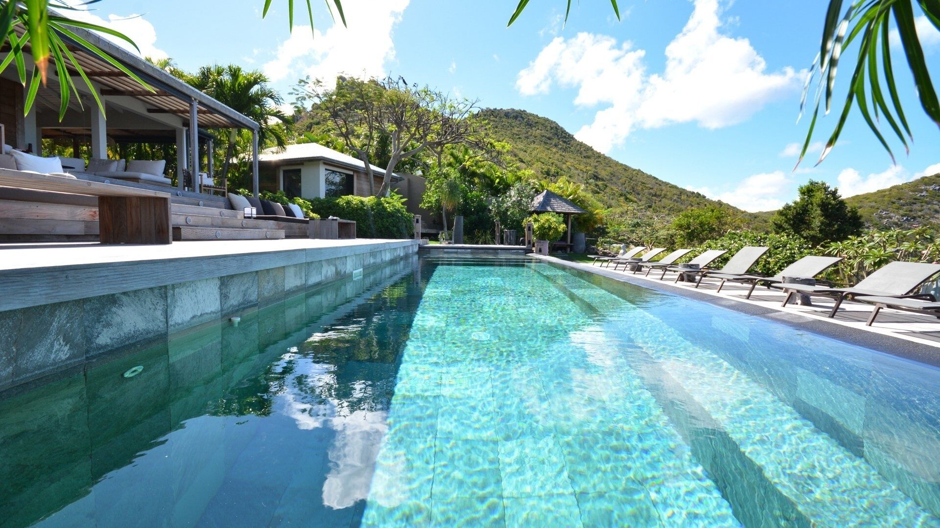 Une piscine aux eaux cristallines, dotée de chaises longues et d’un espace salon abrité, le tout entouré d’une végétation luxuriante et avec une chaîne de montagnes en toile de fond.