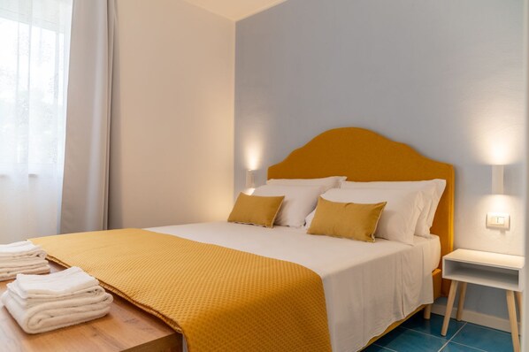 Un lit bien fait, avec une tête de lit jaune, du linge de lit blanc et des oreillers assortis. Une pile de serviettes pliées est posée sur une table en bois, à côté du lit.