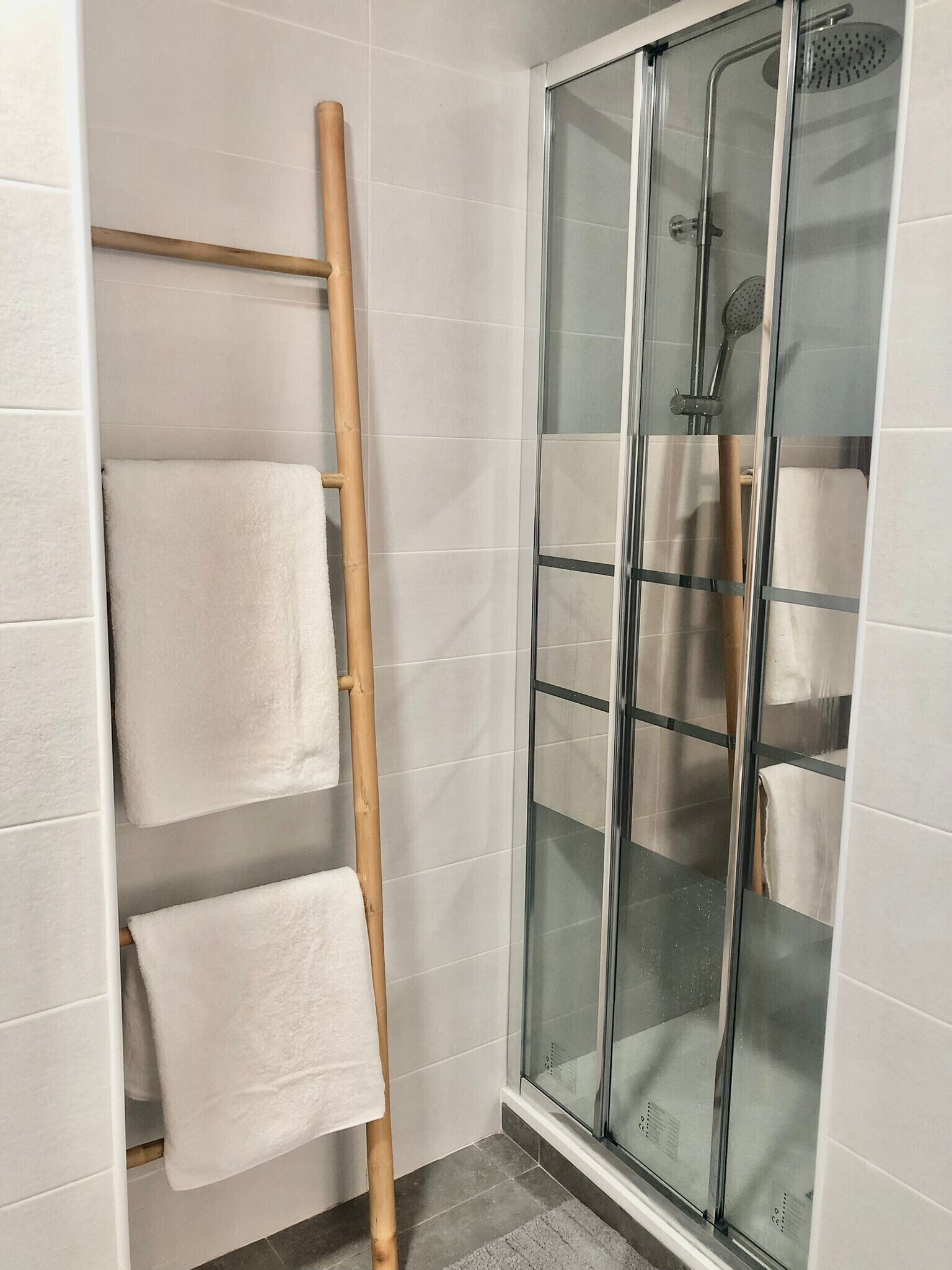 A bathroom with white tiled walls, a glass shower enclosure, and a wooden towel rack with two towels.