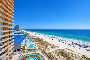 A beachfront hotel with multiple swimming pools, a beach with umbrellas, and a clear blue sky.