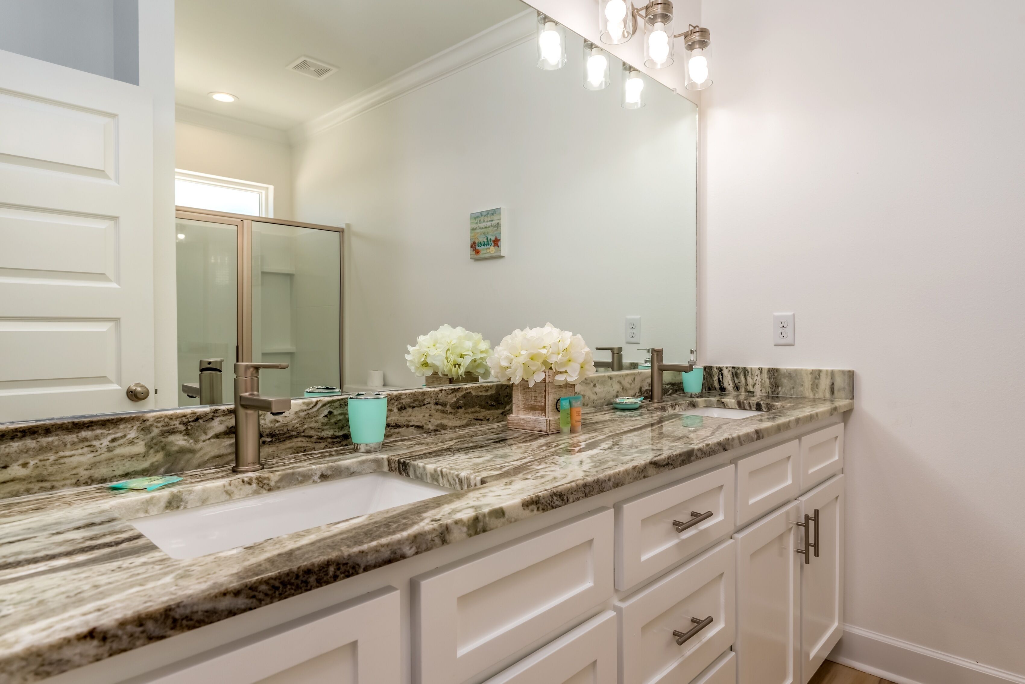 A bathroom with a marble countertop, two sinks, white cabinets, and a large mirror with lights.