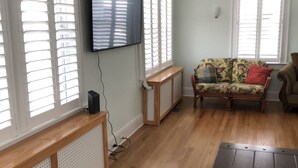 A living room with a wooden floor, a sofa, a TV mounted on the wall, and large windows with blinds. - Bay View Bungalow By The Pier 4 Bedroom Home by RedAwning (Norfolk)
