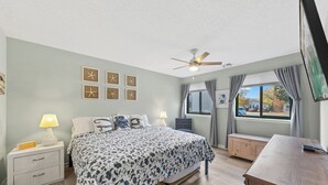 Une chambre à coucher comprenant un lit, des tables de chevet, un bureau, un ventilateur de plafond et une fenêtre avec des rideaux.