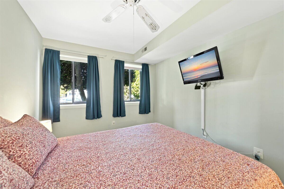 Une chambre à coucher avec un lit, un ventilateur de plafond, un téléviseur fixé au mur et une fenêtre avec des rideaux.