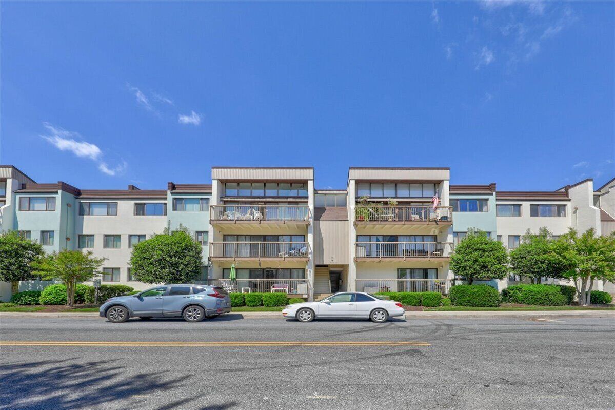 A multi-story apartment building with balconies, a parking lot, and a clear blue sky.