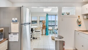 A modern kitchen with a refrigerator, a dining area with a table and chairs, a view through a window, and a small balcony with a chair. - Bliss on the Bay by Seaside Vacations & Sales (Chincoteague Island)